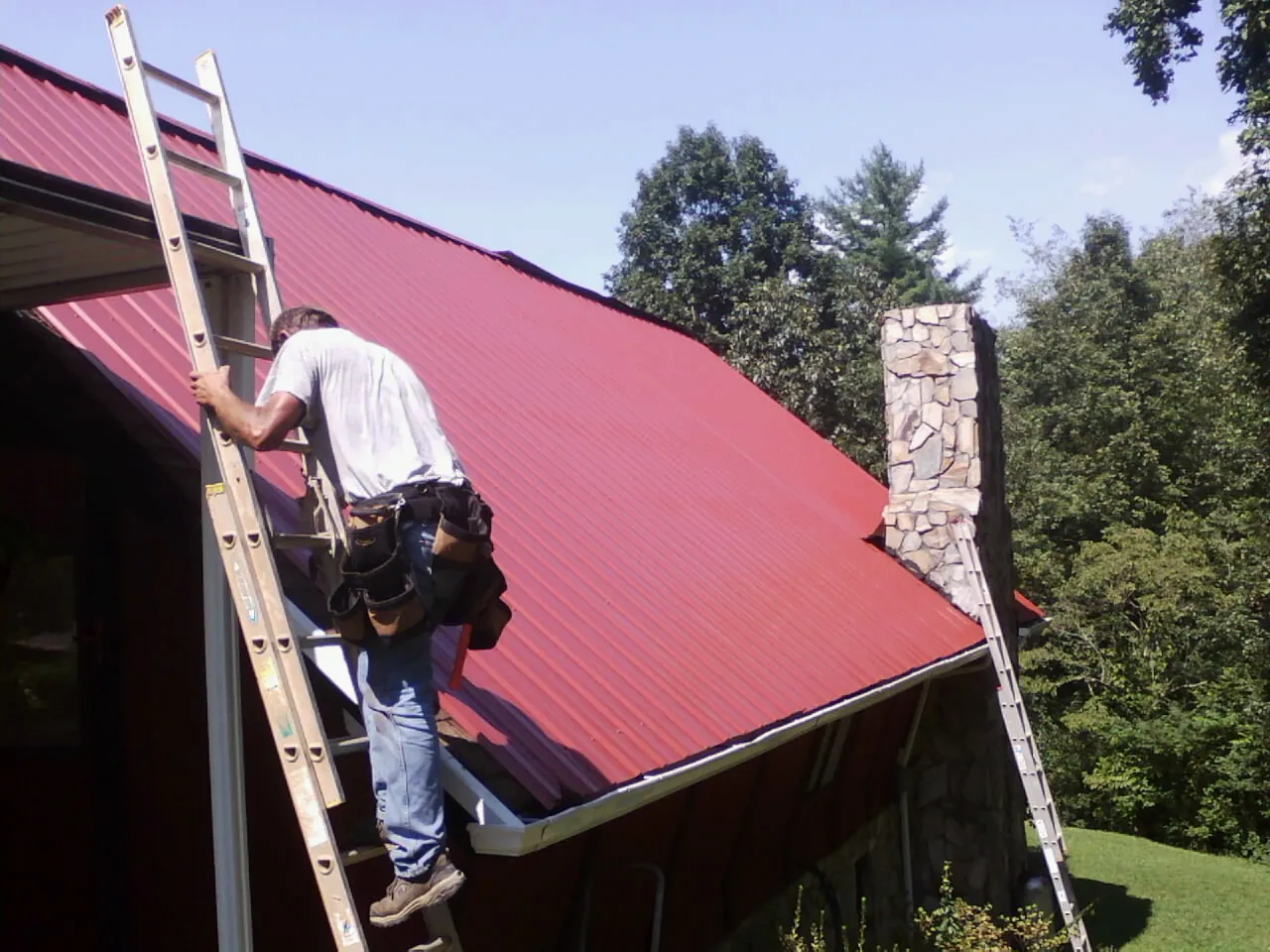 Roofer on a ladder beside a red metal roof and stone chimney — Soffit & Fascia Repair by Roofing Brothers in Balch Springs